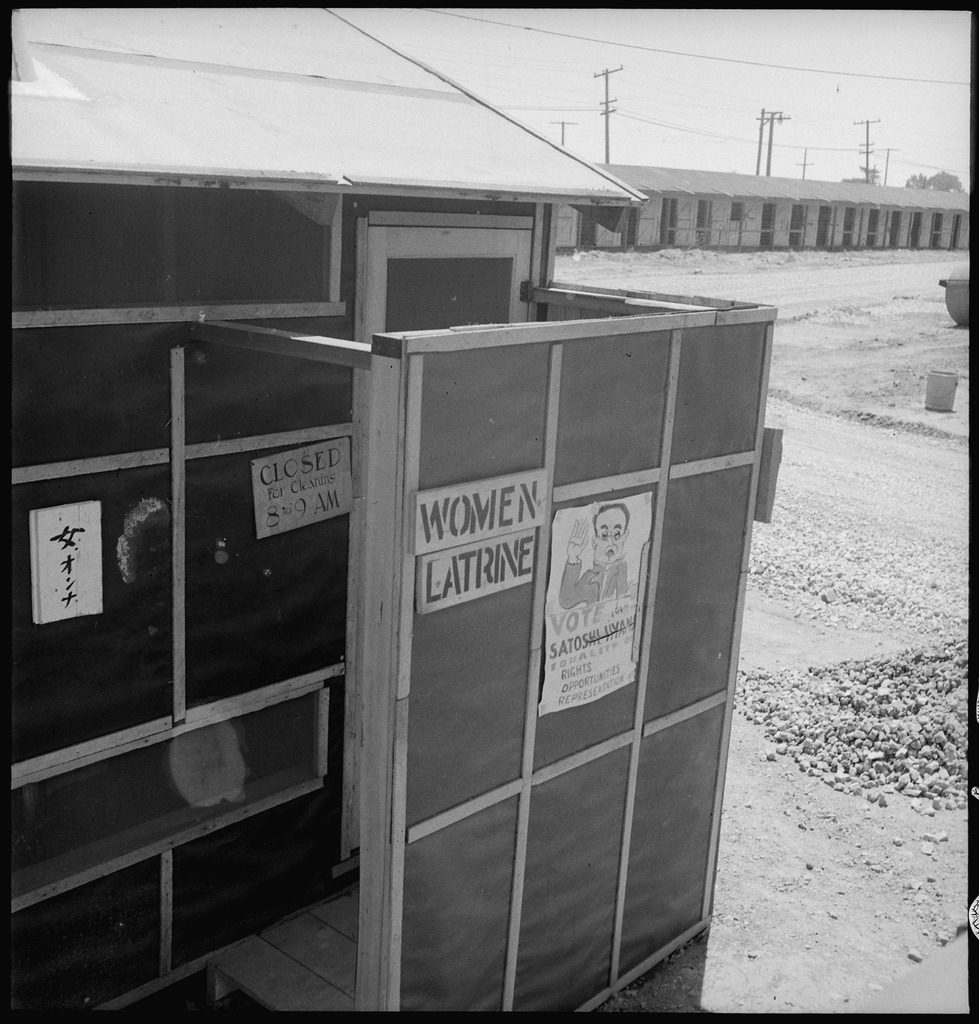 The outside of a latrine at Tanforan Assembly Center. There are signs reading, "Women Latrine" and "Closed for cleaning 8-9am" in English, and "Women" in Japanese posted outside the entrance.