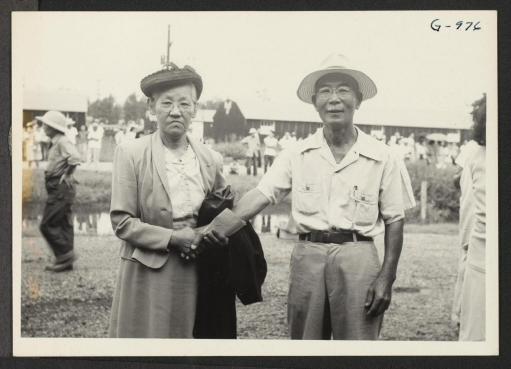 Photo of a Japanese American woman bidding a friend farewell before boarding a train to leave Rohwer concentration camp. She is holding the man's hand while they both look at the camera. Barracks and other people milling about are visible in the background.