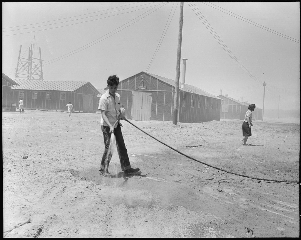 Black and white image of a detainee using a hose. In the background we see imposing electricity poles and barracks.
