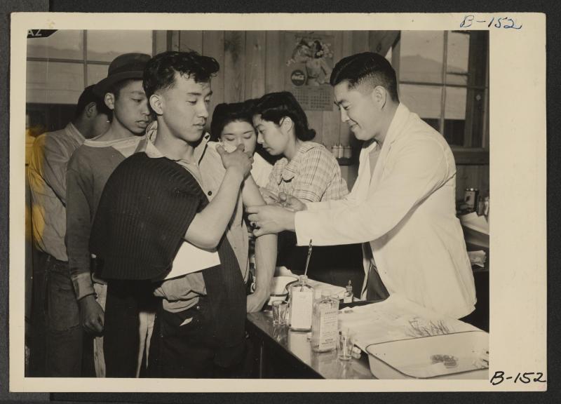 Black and white photo of Nikkei detainees receiving typhoid shots in a line.