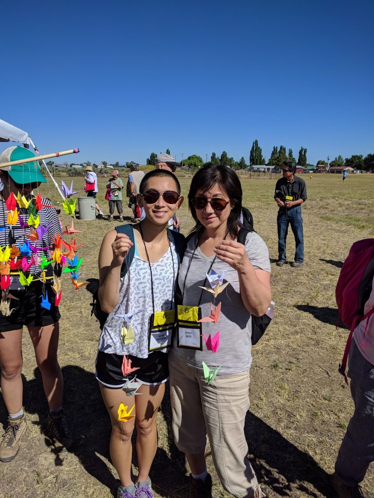 Two Nikkei standing in a large dry grass field amongst a dispersed crowd. Both are holding strands of colorful tsuru.