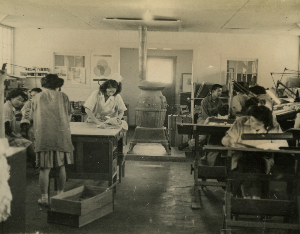 Image of the interior of the Amache Silk Screen Shop. We see several workers attending to different tasks around the shop.