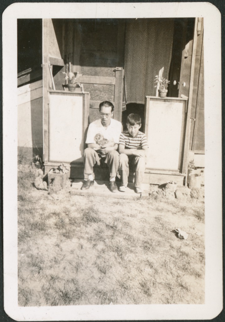A man and a boy sit on a stoop in front of a barrack unit in a Japanese American incarceration camp. There is a curtain hanging in the doorway behind them and potted plants on the stoop. The man is wearing a white shirt, jeans and glasses, and sits on the left holding a small cat. The boy is wearing a striped t-shirt and jeans and sits on the right with his arms resting on his legs.