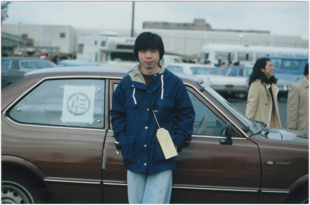 Nikkei person standing with hands in coat pocket in front of a brown car.