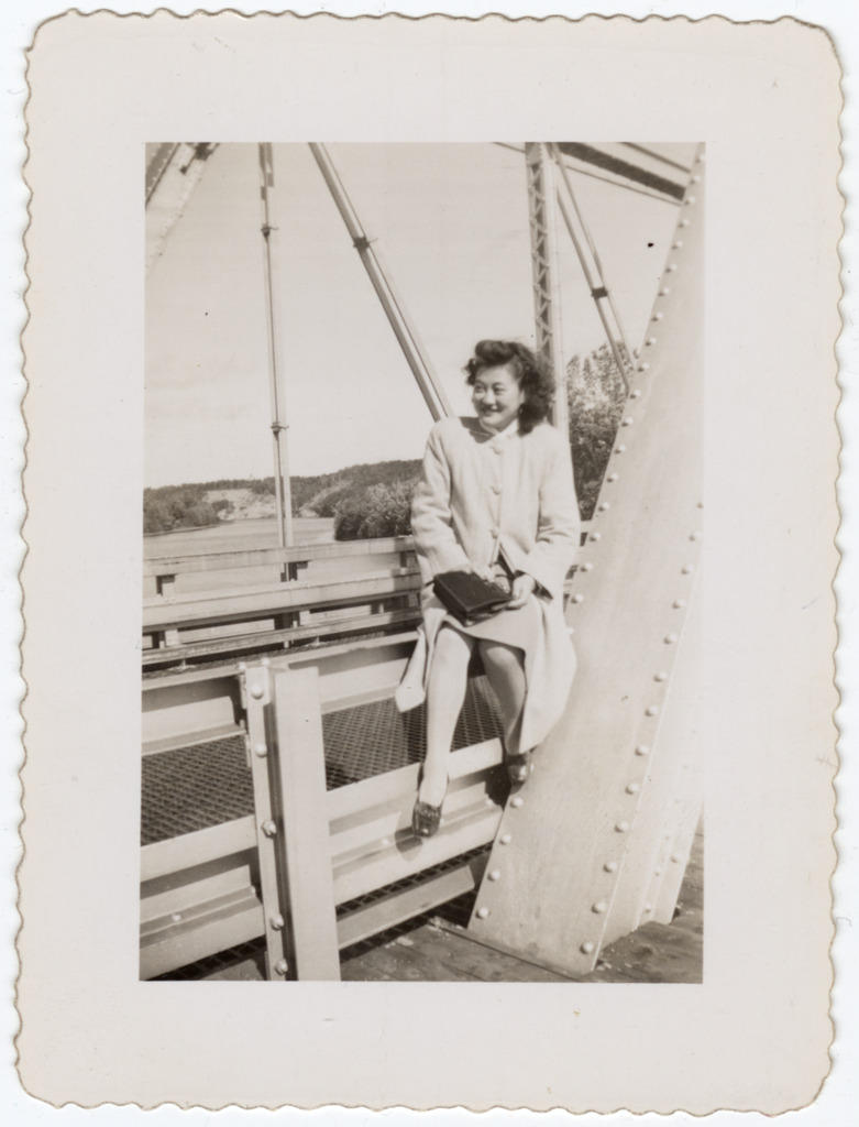 A Japanese American woman sitting on the railing of a steel bridge. She is smiling and wearing a chic coat, and holds a black handbag in her lap.