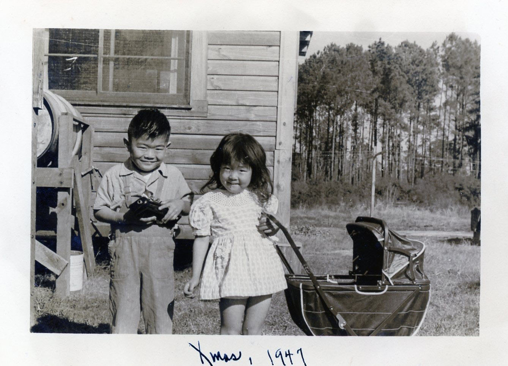 Two Japanese American children posing with toys. A boy on the left holds a small toy and a girl on the right stands next to a toy baby stroller. A house and trees are visible in the background.