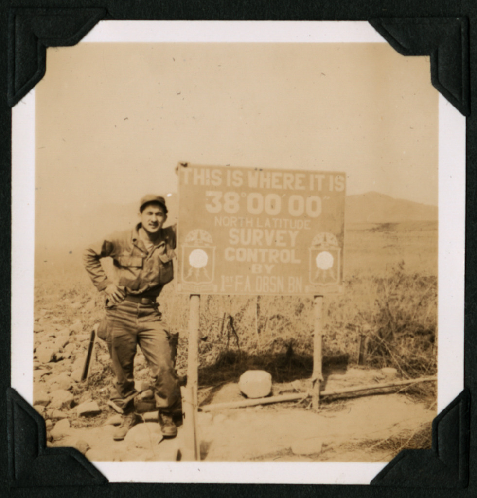 Black and white photo of a uniformed Japanese American soldier posing by a sign at the 38th parallel in Korea, c. 1951.