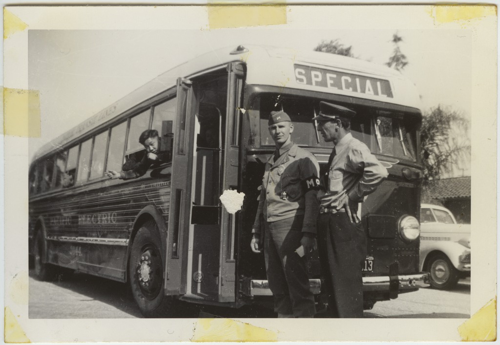 Photo of a bus taking Japanese Americans to an "assembly center" in 1942. A Japanese American boy is leaning out a window on the side of the bus, and a soldier and the bus driver are standing in front of the bus.