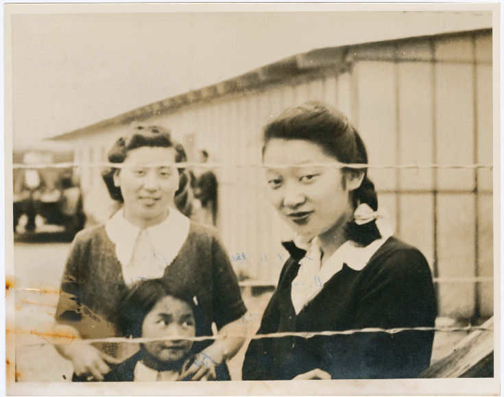 Black and white photo of two Japanese American women and a girl in a WWII concentration camp. Both women are wearing dark sweaters over light blouses and looking at the camera from behind a barbed wire fence. The woman at left stands behind the girl, resting her hands on the girl's shoulders. Visible in the background is a barracks and a car.