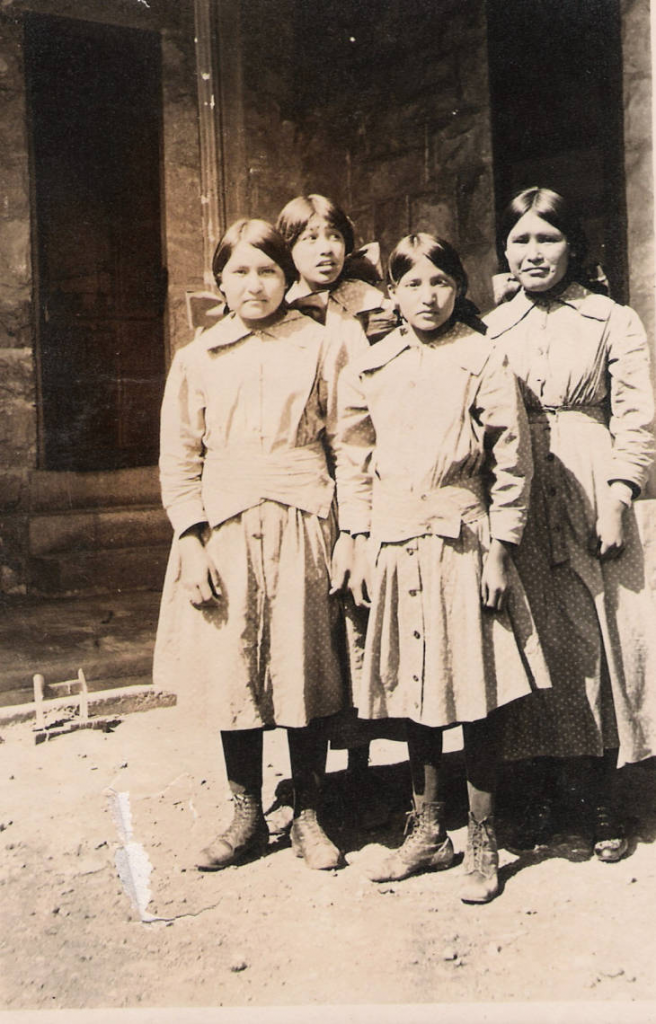 Black and white photo of four Native American girls at the Leupp Boarding School in the early 1920s. They are wearing identical Western dresses and bows in their hair.