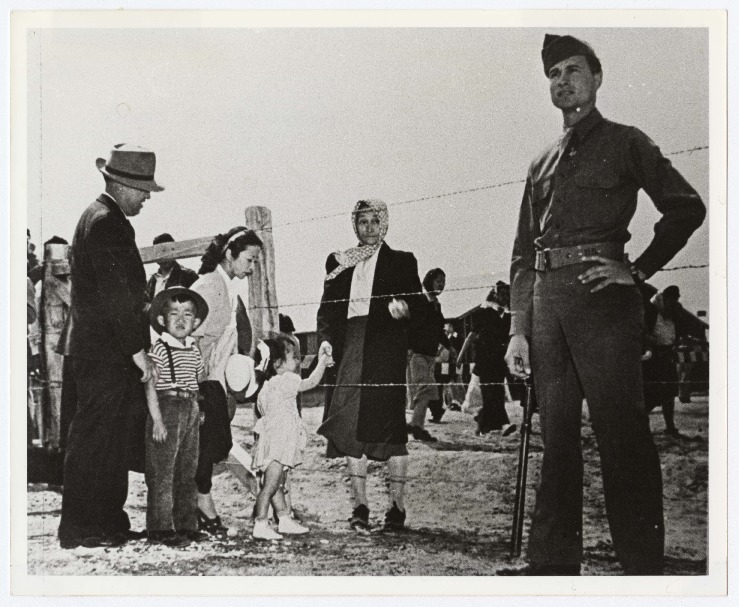 Black and white photo of a family visiting a friend in a Japanese American incarceration camp. A woman wearing a headscarf stands on the inside of a barbed wire fence, holding hands with a young girl on the other side. Next to the girl are her parents and an older brother. An armed sentry stands guard in front of the fence, and other Japanese American inmates and a barracks are visible in the background.