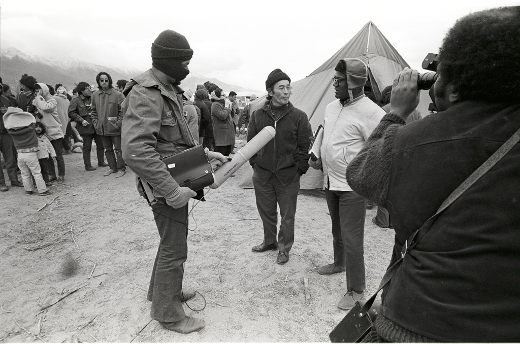 A film crew talking to a pilgrimage attendee. There is one man speaking to the pilgrim while two others hold a camera and a microphone. A tent and other pilgrims are in the background.