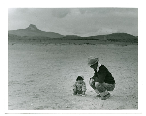 A Japanese American man and child squatting and looking at rocks, with Heart Mountain in the background.
