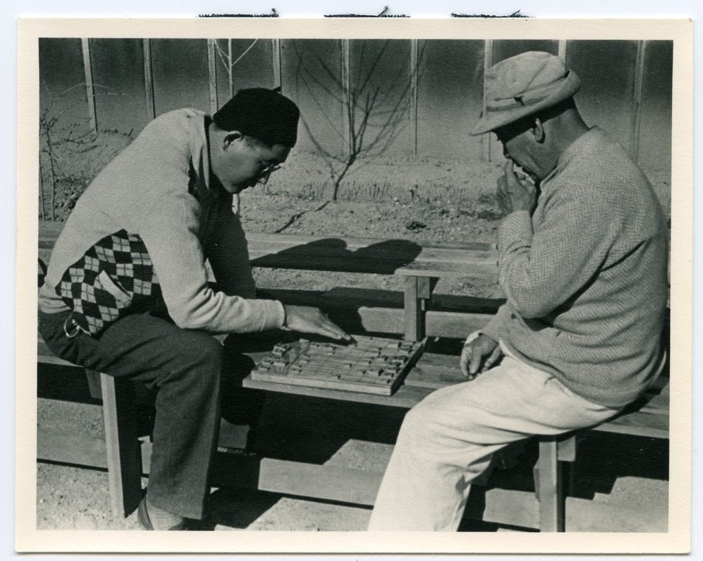 Two men sitting on a bench outside of a barrack building playing a board game. Both men are straddling the bench facing each other with the game between them, and the man at left is leaning over the board making a move.