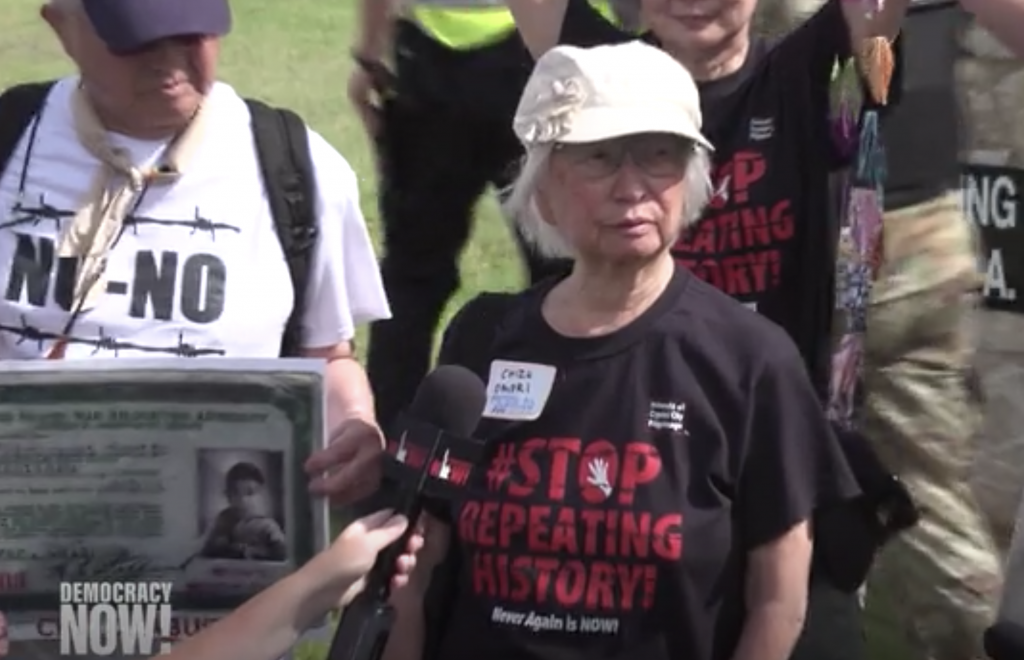 Chizu and others at a protest against the detention of immigrant and refugee children. Chizu is wearing a black t-shirt with "Stop Repeating History" in red letters and speaking into a microphone.
