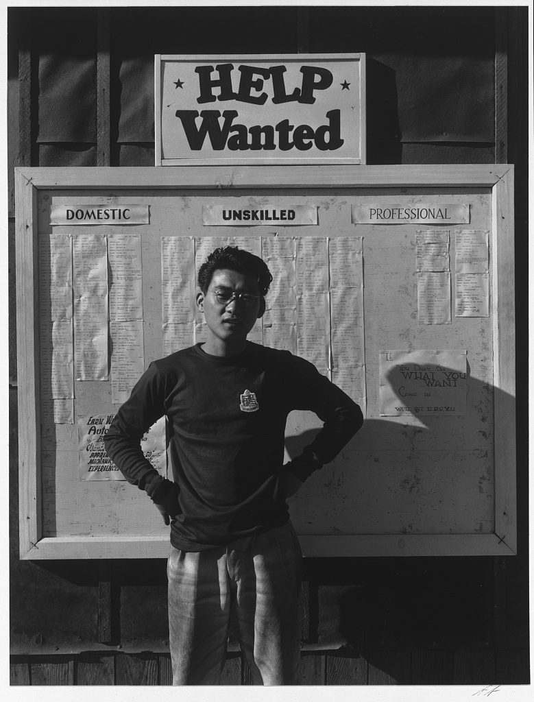 A young Japanese American man standing in front of a board with job postings and a large "Help Wanted" sign. He is standing with his hands on his hips and looking directly at the camera.