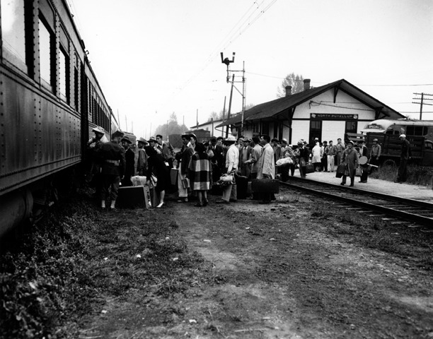 A crowd of Japanese Americans at a train depot, boarding a train that will take them to Manzanar.